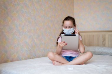 Koruyucu maskeli, tabletli beyaz bir kız evde kendini tecrit modunda yatağın üzerinde oturuyor..