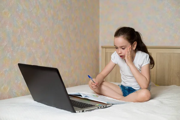 遠隔オンライン教育 デジタルタブレットノートパソコンで自宅で勉強し 学校の宿題をしている女子高生 トレーニング本と一緒にベッドに座って — ストック写真