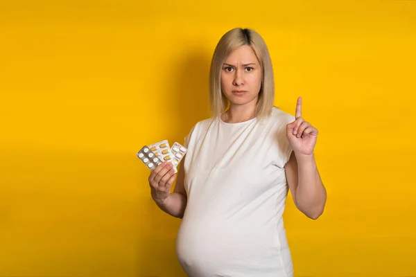 serious pregnant woman, blonde warns by holding her index finger up, holds in his hands the tablets on a yellow background. safe medications for pregnancy.