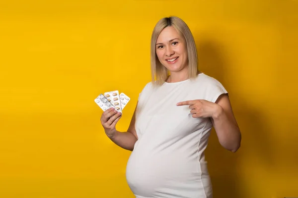 happy pregnant woman, blonde points her finger at the pills on a yellow background. safe medications for pregnancy.