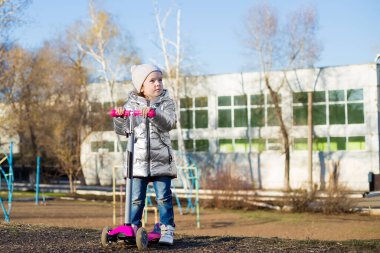 Güneşli bir bahar gününde parkta scooter süren küçük bir kız. Çocuklar için aktif eğlence ve açık hava sporu.