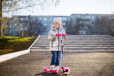 Güneşli bir bahar gününde parkta scooter süren küçük bir kız. Çocuklar için aktif eğlence ve açık hava sporu.