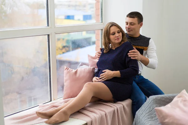 Een jonge zwangere vrouw zit voor een groot raam met haar gelukkige man die haar omhelst. het concept van een jong gezin — Stockfoto