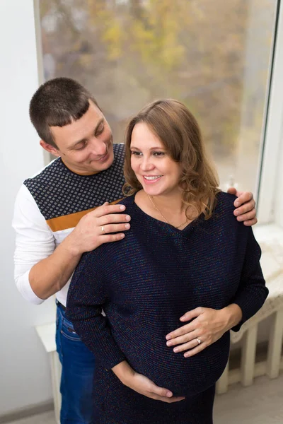 Μια νεαρή έγκυος γυναίκα κάθεται σε ένα μεγάλο παράθυρο με τον ευτυχισμένο σύζυγό της να την αγκαλιάζει. η έννοια της νέας οικογένειας — Φωτογραφία Αρχείου
