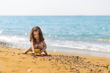 Denizin arka planında tropik plajda kumla oynayan tatlı küçük bir kız. Aile tatili kavramı.