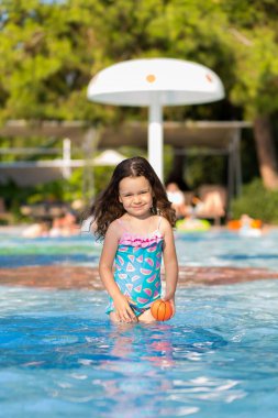 Güneşli bir yaz gününde, parlak bir mayo giymiş, havuzda top oynayan mutlu küçük bir kız..