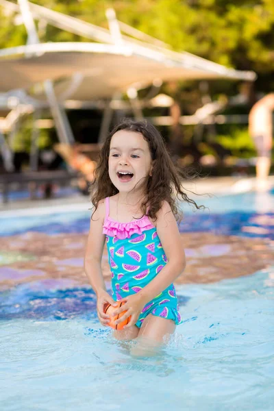 Güneşli bir yaz gününde, parlak bir mayo giymiş, havuzda top oynayan mutlu küçük bir kız..