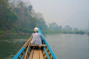 Yeşil ormandaki vahşi bir tropikal nehirde gezinen küçük ahşap bir tekne. Teknenin önünde bir kadın oturuyor ve manzarayı huzur içinde seyrediyor. Mavi kano doğayı keşfediyor..