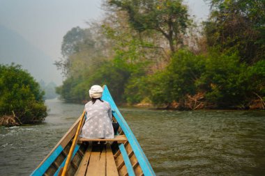 Yeşil ormandaki vahşi bir tropikal nehirde gezinen küçük ahşap bir tekne. Teknenin önünde bir kadın oturuyor ve manzarayı huzur içinde seyrediyor. Mavi kano doğayı keşfediyor..