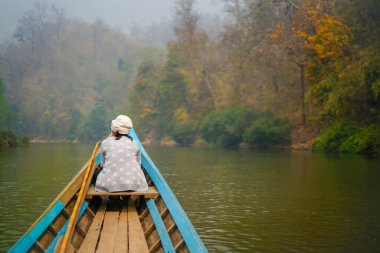 Yeşil ormandaki vahşi bir tropikal nehirde gezinen küçük ahşap bir tekne. Teknenin önünde bir kadın oturuyor ve manzarayı huzur içinde seyrediyor. Mavi kano doğayı keşfediyor..