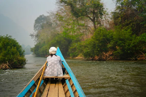 Yeşil ormandaki vahşi bir tropikal nehirde gezinen küçük ahşap bir tekne. Teknenin önünde bir kadın oturuyor ve manzarayı huzur içinde seyrediyor. Mavi kano doğayı keşfediyor..