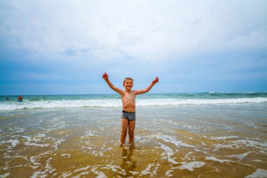 Arkasında dalgalar ve deniz olan neşeli bir çocuk. Neşeli ve neşeli çocuk deniz kıyısında eğleniyor. 