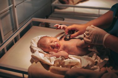 Doktor çocuk doktoru klinikte bebeği muayene ediyor, yeni doğmuş bebek yatakta ağlıyor.