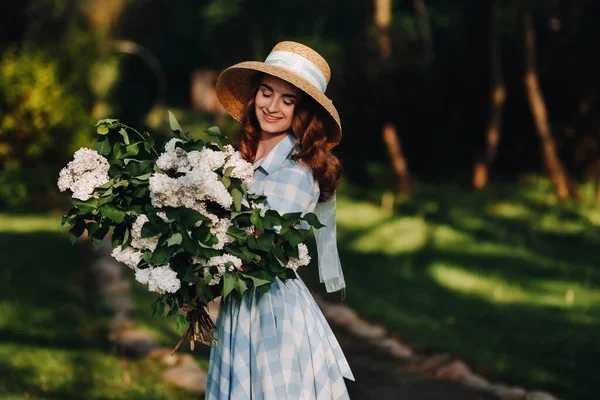 Eine stylische Frau mit Strohhut posiert auf fliederfarbenen Blumen in einem sonnigen Frühlingspark. Ruhiges Porträt eines schönen Mädchens, das mit einem Strauß Flieder im Frühlingsgarten steht. — Stockfoto