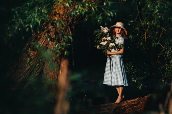 Uma mulher elegante com um chapéu de palha e pés nus posa em uma árvore caída com um buquê de lilases em um Sunny Spring Park. Um retrato tranquilo de uma linda garota de pé em um lilás roxo — Fotografia de Stock