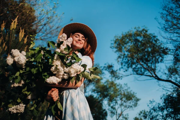 ストローハットのスタイリッシュな女性とストローバッグは、サニースプリングパークで白いライラックの花束でポーズをとっています。春の庭で紫のライラックブッシュに立つ美しい少女の静かな肖像画. — ストック写真