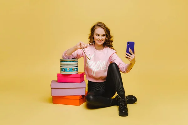 Una joven feliz con cajas de papel coloridas después de ir de compras, sentado y tomando selfies aislados en un fondo naranja Studio. Ventas estacionales, compras, gastar dinero en regalos — Foto de Stock