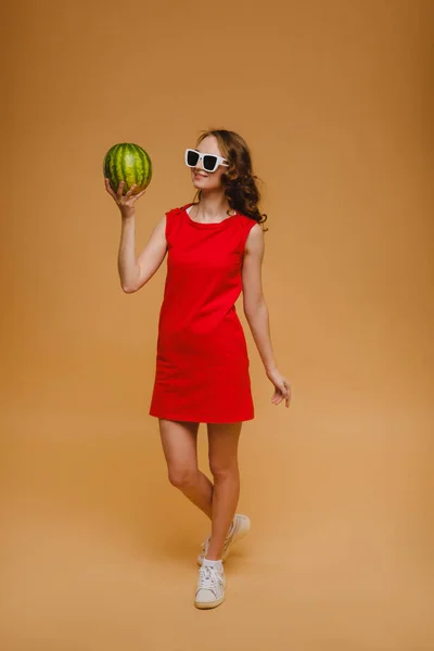 Una hermosa chica en gafas y un vestido rojo sostiene una sandía en sus manos — Foto de Stock