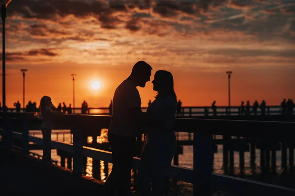 Silhueta retrato de um belo casal recém-casado. Um homem amoroso abraça uma menina ao pôr-do-sol, contra o fundo do mar em pé no cais . — Fotografia de Stock