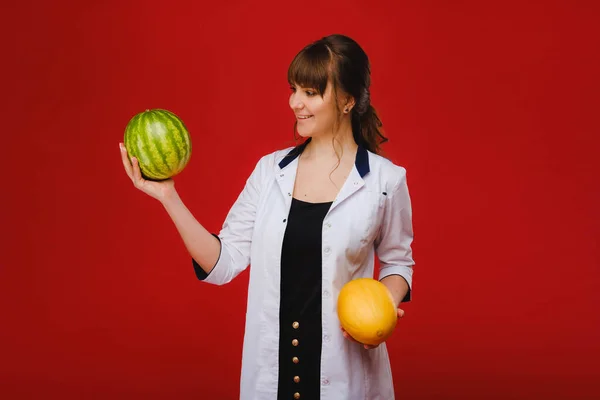 Una enfermera doctora con una bata blanca con fruta en las manos posa sobre un fondo rojo, melón, sandía , — Foto de Stock