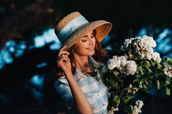 ストローハットのスタイリッシュな女性は サニースプリングパークでライラックの花束でポーズをとっています 春の庭に立つ美しい少女の静かな肖像画 — ストック写真