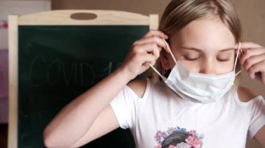 Evdeki küçük bir kız COVID-19 yazısının arkasında tıbbi maske takıyor.