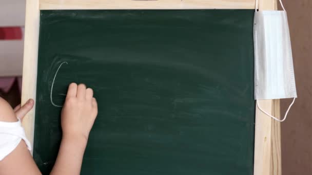 Een klein meisje thuis met een masker schrijft een vraag met krijt op het schoolbord: God zegene u — Stockvideo