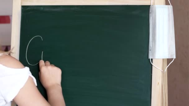 Een klein meisje met een masker schrijft het woord COVID-19 op het schoolbord met krijt. — Stockvideo