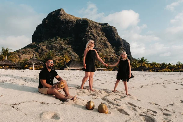 Una familia elegante en ropa negra con cocos en sus manos en la playa de la isla de Mauricio. Hermosa familia en la isla de Mauricio en el contexto del monte Le Morne Brabant — Foto de Stock