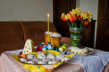 Paskalya masası düzeni. Masanın üzerinde üç çeşit Paskalya yumurtası, yanan bir mum ve bir buket çiçek var.