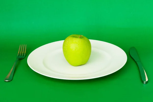Health care diet concept green apple in white plate with knife and fork isolated in green background. — Stock Photo, Image