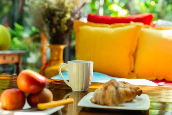 Mugg kaffe med äpple och croissant på träbord med färgglad soffa i huset. — Stockfoto