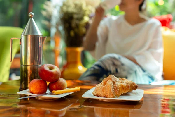 Mujer borrosa disfruta del café de la mañana con croissants y manzana en la mesa de madera . —  Fotos de Stock