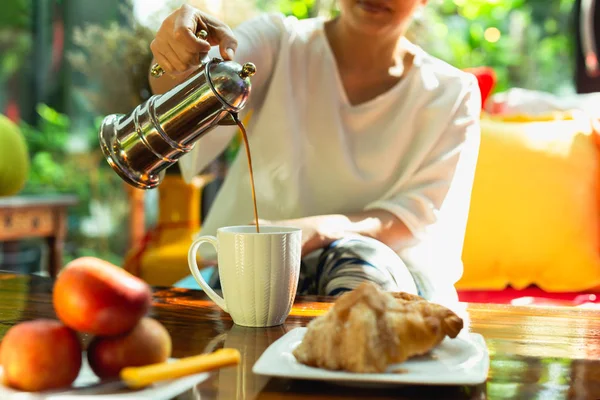 テーブルの上に食べ物のあるコーヒーメーカーからカップにコーヒーを注ぐ女性. — ストック写真