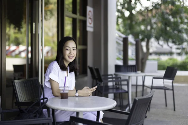 Feliz mujer sonriente con café helado y teléfono celular sentado en la terraza de la cafetería al aire libre . —  Fotos de Stock