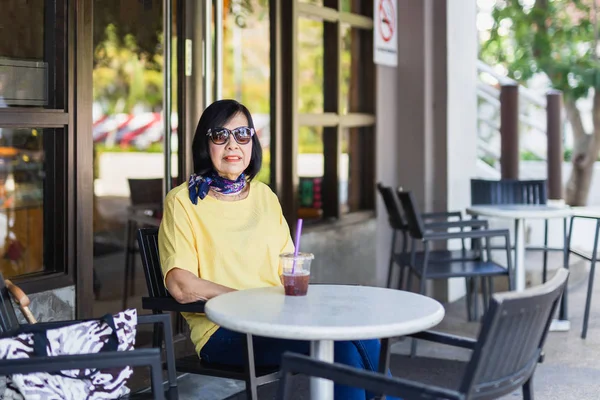 Old woman with sun glasses seated at a coffee shop in the shade.