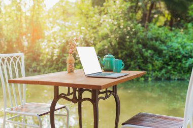 Kanalın kenarındaki ahşap masada çay fincanı ve gül meyveli dizüstü bilgisayar.