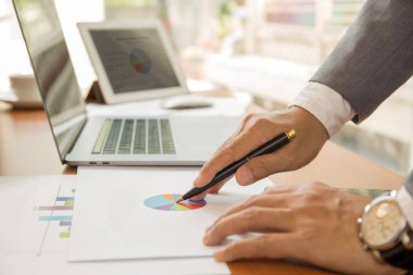 İşadamı elinde kalemle masadaki dizüstü bilgisayarlar ve tabletlerle finansal diyagramı gösteriyor.
