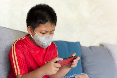 Koruyucu maskeli Asyalı çocuk evde cep telefonuyla oyun oynuyor.