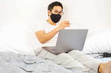 Yatakta koruyucu maskeli bir adam termometre koronavirüs karantinasına bakıyor.