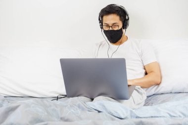 Koruyucu maskeli ve kulaklıklı bir adam evde dizüstü bilgisayarda çalışıyor.