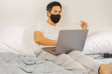 Yatakta koruyucu maskeli bir adam termometre koronavirüs karantinasına bakıyor.