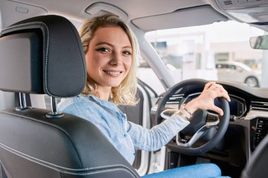 Araba galerisindeki güzel kadın, kameraya bakıyor ve yeni arabanın içinde otururken gülümsüyor. 