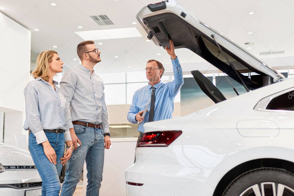 Sales manager helping young couple choose a new car by showing the size and interior of a trunk