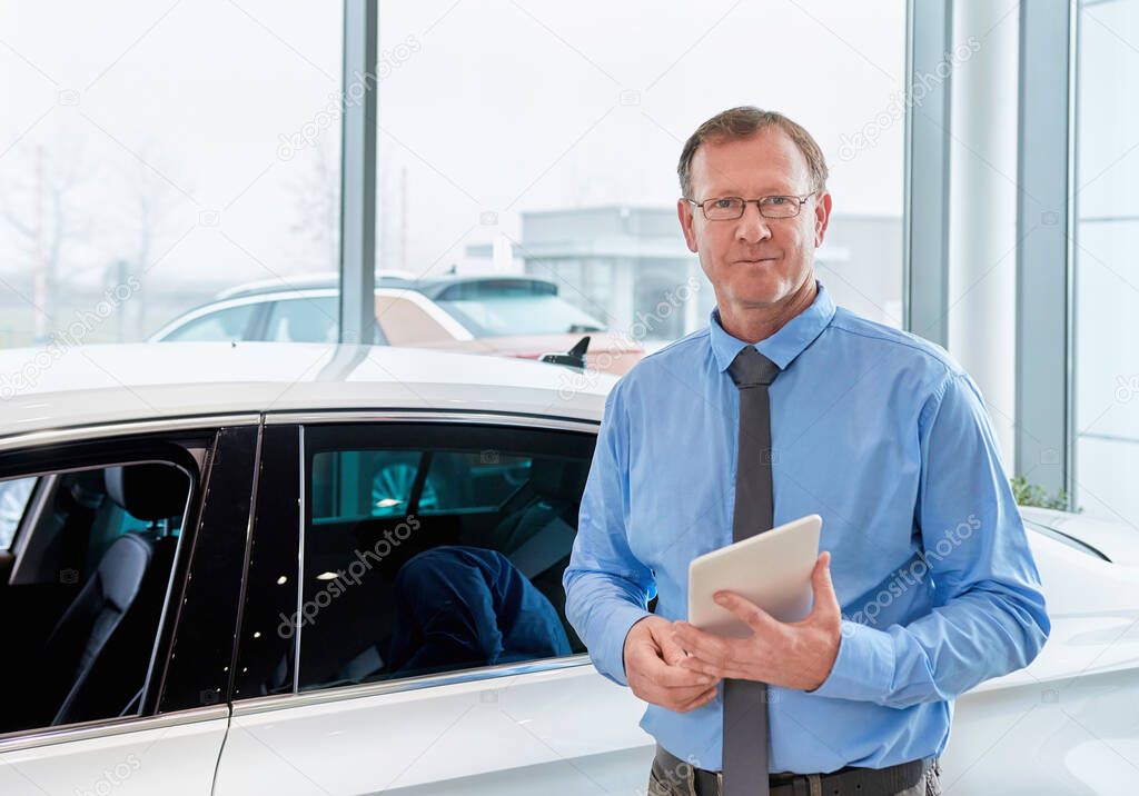 Car salesman posing in front of a vehicle at car dealership, holding tablet