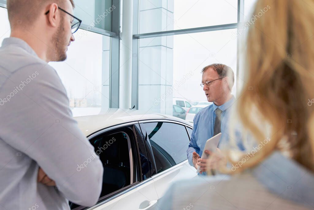 Car dealer helping young couple choose a new car by showing the advantages of the vehicle to the customers