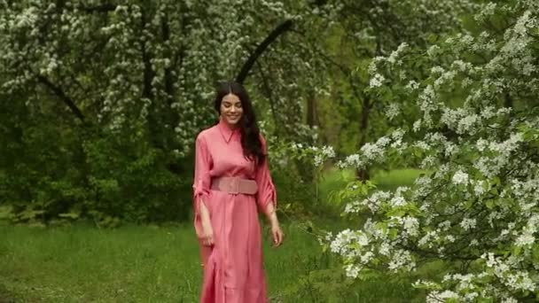 Gyönyörű divatos nő rózsaszín ruhában sétál a virágzó kertben — Stock videók