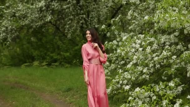 Hermosa chica morena elegante con el pelo largo en vestido rosa está caminando en el jardín floreciente — Vídeo de stock