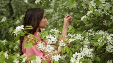 Pembe elbiseli, uzun siyah saçlı güzel moda kızı çiçek açan ormanda beyaz çiçeklere bakıyor.