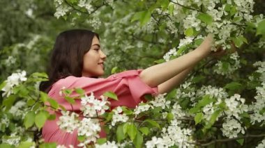 Pembe elbiseli güzel, modaya uygun bir kız beyaz taç yaprakları topluyor ve çiçek açan ormandaki ellerden üflüyor.